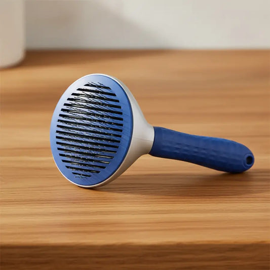 A blue and white pet grooming brush with a flexible bristle head and a curved handle, resting on a wooden tabletop against a neutral beige background.