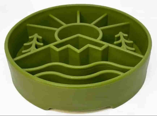 An olive green silicone pet slow feeder bowl with a landscape design featuring a sun, mountains, trees, and wavy water lines, shown on a plain white background.