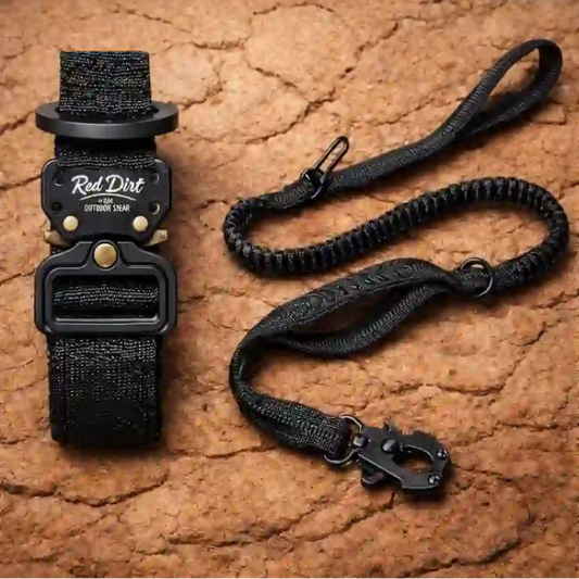 A black tactical belt with a matching paracord lanyard strap, displayed on a textured brown stone background. The buckle is black with brass hardware and features the 'Red Dirt' brand logo.